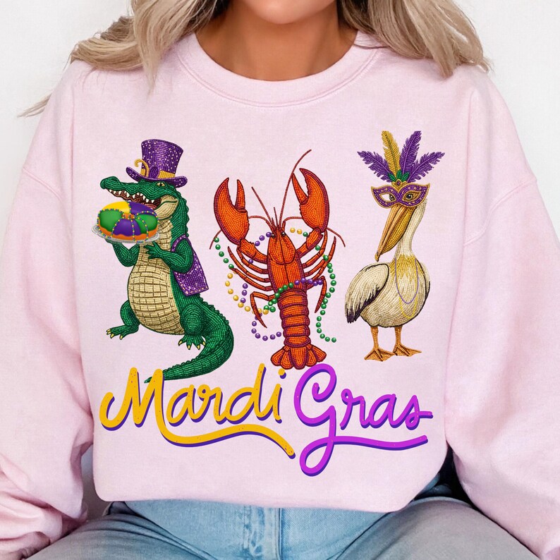 May include: A light pink sweatshirt featuring a colorful Mardi Gras design. The design includes a green alligator wearing a purple hat, an orange lobster, and a white pelican wearing a mask. The words "Mardi Gras" are written in yellow and purple.