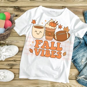 Puede incluir: Camiseta blanca con un diseño retro que presenta una taza de café, un café con leche de calabaza y un balón de fútbol americano. Las palabras "FALL VIBES" están en naranja, con hojas de otoño y estrellas.