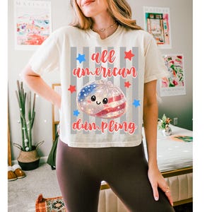 May include: Cream-colored t-shirt with the text "all american dumpling" in red script. The design features a dumpling with an American flag design, surrounded by red and blue stars. The shirt is a casual, comfortable style.
