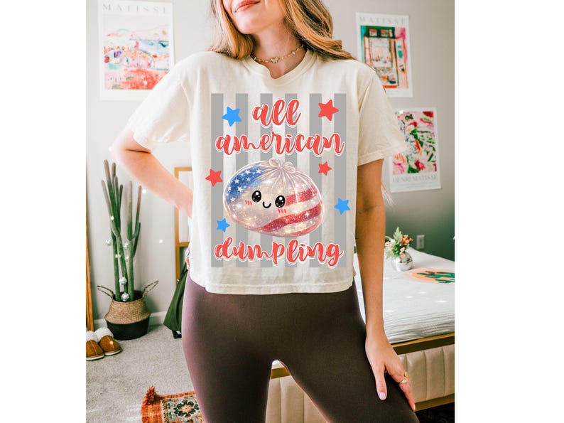 May include: Cream-colored t-shirt with the text "all american dumpling" in red script. The design features a dumpling with an American flag design, surrounded by red and blue stars. The shirt is a casual, comfortable style.