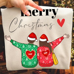 Könnte beinhalten: Beiges Sweatshirt mit den Worten „Merry Christmas“ und einem roten Herz. Das Sweatshirt zeigt zwei Figuren mit Weihnachtsmützen und Weihnachtspullovern mit personalisierten Herzdesigns. Eine Hand zeigt auf das Sweatshirt.