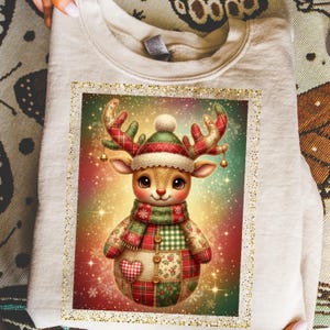 May include: A beige sweatshirt featuring a colorful, cartoon-style reindeer design. The reindeer wears a hat, scarf, and patchwork outfit in red, green, and white. The image has a gold glitter border and a festive, sparkling background.