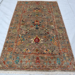 Könnte beinhalten: Ein rechteckiger Teppich mit einem gedämpften braunen Hintergrund und einem detaillierten Blumenmuster in Rot-, Blau-, Grün-, Orange- und Gelbtönen. Der Teppich hat eine dekorative Bordüre und kurze weiße Fransen an den oberen und unteren Kanten.