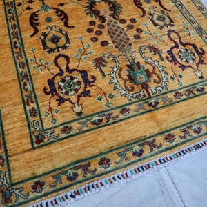 Puede incluir: Una alfombra rectangular con un fondo amarillo dorado vibrante y motivos florales y botánicos intrincados en tonos azul profundo, rojo oscuro y verde azulado. La alfombra está enmarcada por un borde decorativo y flecos multicolores.