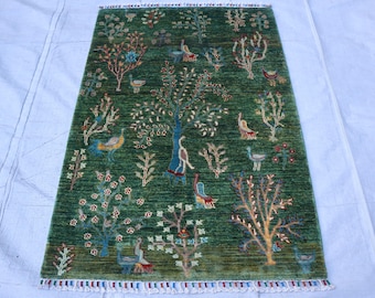 May include: A rectangular green rug with a detailed pattern of trees, birds, and floral elements. The rug has a fringe border with blue, red, and white accents. The design features various colors, including blue, red, and brown.