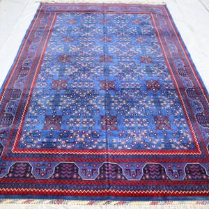 May include: A blue rectangular rug with a detailed pattern. The rug has a central blue field with repeating geometric designs in red, purple, and white. A red border with intricate patterns frames the rug, adding a decorative element.