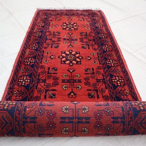 May include: A rolled-up red rug with a detailed geometric pattern in navy blue and cream. The rug is partially unrolled, revealing the intricate design. The rug's fringe is visible at the top. This rug is suitable for home decor.