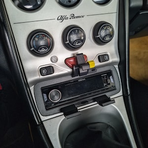 May include: Car dashboard with silver trim, featuring gauges, dials, and a radio. The Alfa Romeo logo is displayed. Gauges indicate fuel, temperature, and other vehicle data. A black radio is installed in the centre console.