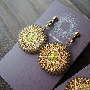 May include: A pair of gold clip-on earrings with a yellow, white and orange circular design. The earrings are displayed on a grey card with the text "okamotohama" printed on it.