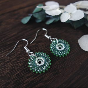 Può includere: Un paio di orecchini circolari avvolti in filo verde e bianco con ganci d'argento. Gli orecchini hanno un design delicato e intricato.