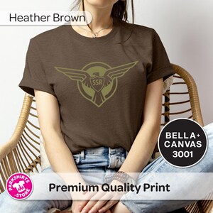 May include: Heather brown t-shirt with an olive green emblem of an eagle with outstretched wings and the letters "SSR". The shirt is made of Bella+Canvas 3001 material.