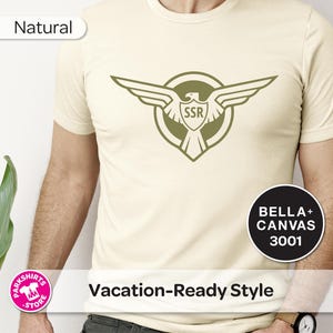 May include: A cream-coloured t-shirt featuring a green eagle emblem with "SSR" inside a shield. The shirt also displays the text "Vacation-Ready Style" and "Natural". Made from Bella+Canvas 3001 material.