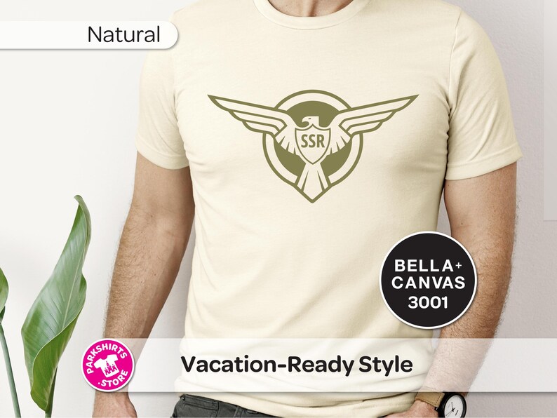 May include: A cream-coloured t-shirt featuring a green eagle emblem with "SSR" inside a shield. The shirt also displays the text "Vacation-Ready Style" and "Natural". Made from Bella+Canvas 3001 material.