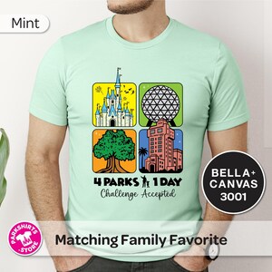 Puede incluir: Camiseta verde menta con un gr&aacute;fico colorido que presenta cuatro iconos de parques tem&aacute;ticos. El dise&ntilde;o incluye un castillo, una esfera geod&eacute;sica, un &aacute;rbol y una torre. El texto dice "4 Parks 1 Day Challenge Accepted".