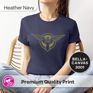 May include: Navy blue t-shirt with a gold emblem of wings and the letters "SSR". The shirt is made of Bella+Canvas 3001 material. The words "Premium Quality Print" are also visible.