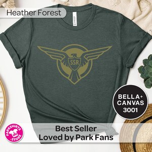 May include: A dark green t-shirt with a gold emblem featuring an eagle and the letters "SSR". The shirt has rolled-up sleeves and the text "Heather Forest" at the top. The text "Best Seller Loved by Park Fans" is at the bottom.