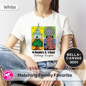 Puede incluir: Camiseta blanca con un gr&aacute;fico colorido que presenta cuatro iconos de parques tem&aacute;ticos. El dise&ntilde;o incluye un castillo, una esfera geod&eacute;sica, un &aacute;rbol y una torre, con el texto "4 PARKS 1 DAY Challenge Accepted". La camiseta est&aacute; etiquetada como "BELLA+ CANVAS 3001".