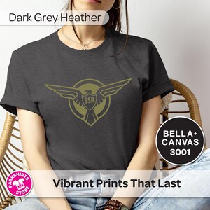 May include: Dark grey heather t-shirt with a gold emblem of wings and the letters "SSR" inside a shield. The shirt is labelled "BELLA + CANVAS 3001" and has the text "Vibrant Prints That Last".