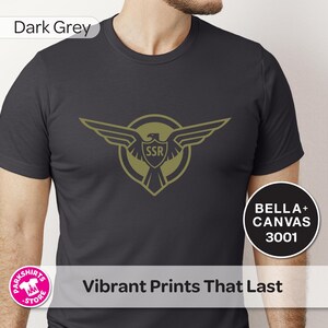 May include: Dark grey t-shirt with a gold eagle emblem and the letters "SSR". The shirt displays the text "Vibrant Prints That Last" and "BELLA + CANVAS 3001".