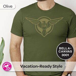 May include: Olive green t-shirt featuring an eagle emblem with "SSR" inside a shield. The shirt also has the text "Vacation-Ready Style" and the brand "BELLA + CANVAS 3001".