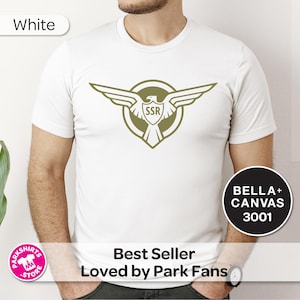 May include: White t-shirt with a green winged emblem and "SSR" within a shield. The shirt is a Bella+Canvas 3001. Text includes "Best Seller Loved by Park Fans". The shirt is a popular item.