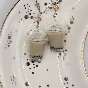 May include: A pair of dangle earrings featuring miniature clear plastic cups filled with a light brown liquid and black spheres at the bottom, resembling bubble tea. Each earring has a silver-toned metal hook and a small, clear faceted bead.
