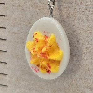 May include: Novelty earrings with a devilled egg design. Each earring has a white egg shape with a yellow and red filling, suspended from a silver-tone hook with a yellow crystal bead.