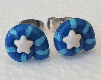 May include: A pair of stud earrings shaped like spiral seashells in shades of dark and light blue, each featuring a small, centered white star. The earrings have silver-toned metal posts and backings, showcasing a glossy, smooth finish.