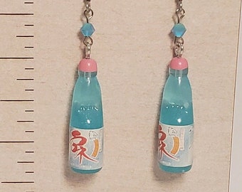May include: A pair of dangle earrings featuring miniature blue soda bottles with pink caps and white labels with red Japanese text. The earrings hang from silver-toned hooks with small blue beads. A ruler shows the earrings are 1.5 inches long.