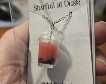 May include: A silver chain necklace with a pink bubble tea charm. The charm is made of clear plastic with a pink liquid inside and black boba pearls. The necklace is packaged in a clear plastic box with a white card that says "Starfall at Dusk" and "Nickel-free Jewelry" with the website address "www.starfallatdusk.com".