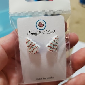 Peut inclure: Une paire de boucles d'oreilles à clous blanches en forme de sapin de Noël avec des motifs de lignes ondulées rouges et vertes. Les boucles sont fixées sur une carte de présentation blanche marquée Starfall at Dusk avec un logo de beignet.