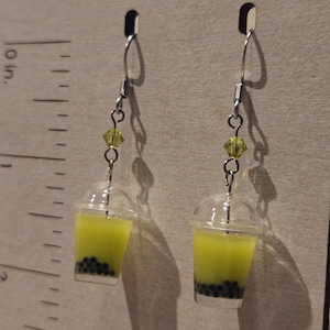 May include: A pair of dangle earrings featuring miniature clear plastic cups filled with yellow liquid and black spheres, resembling bubble tea. Each earring has a lime green faceted bead on a silver-tone hook, measuring approximately 1.5 inches.