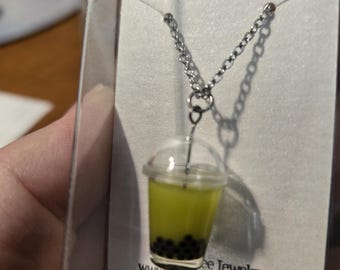 May include: A silver chain necklace with a green and black boba tea charm. The charm is in a clear plastic cup with a straw. The necklace is displayed on a white card with the text "Starfall at Dusk" and a website address.