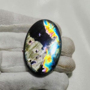 May include: An oval-shaped labradorite gemstone. The stone has a dark base color with iridescent flashes of blue, yellow, and green. The polished surface reflects light, showcasing a play of colors. The gemstone is held in a white fabric.