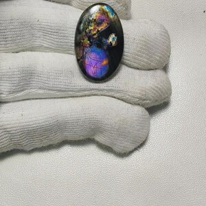 May include: Oval-shaped gemstone with a polished surface. The stone displays a vibrant array of colors, including purple, blue, yellow, and green, creating a shimmering effect. The gemstone is held in a white glove, highlighting its iridescent qualities.