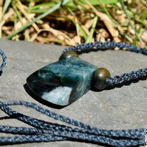 May include: A necklace featuring a polished, rectangular green and white stone pendant suspended on a dark blue braided cord. Two small, round, dark brown wooden beads are threaded onto the cord on either side of the stone. The logo MAYAN MOUNTAIN is visible in the bottom right corner.