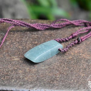 May include: A rectangular, pale green stone pendant with subtle white speckles, suspended from a dark purple braided cord necklace. The cord features adjustable sliding knots and a smooth, polished finish on the stone surface, creating a simple pendant.
