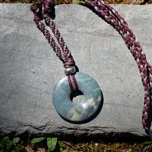 May include: A blue-green stone pendant with a central hole, suspended from a braided cord necklace. The cord is a mix of burgundy and gray strands. The pendant rests on a gray stone surface, with the Mayan Mountain logo visible.