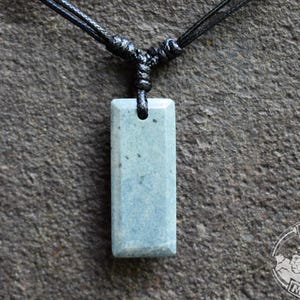 May include: A rectangular, light blue pendant necklace with a black cord. The pendant is made of a stone-like material and has a small hole at the top. The background is a textured, dark gray surface. The Mayan Mountain logo is in the bottom right corner.