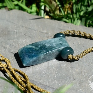 May include: A rectangular, mottled dark green stone pendant with a smooth, polished finish. The pendant is attached to a tan, braided cord necklace featuring two small, round, dark green stone beads flanking the central rectangular piece.