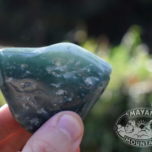 May include: A green and white agate stone with a rough, natural texture. The stone is held in a hand, and the background is blurred.