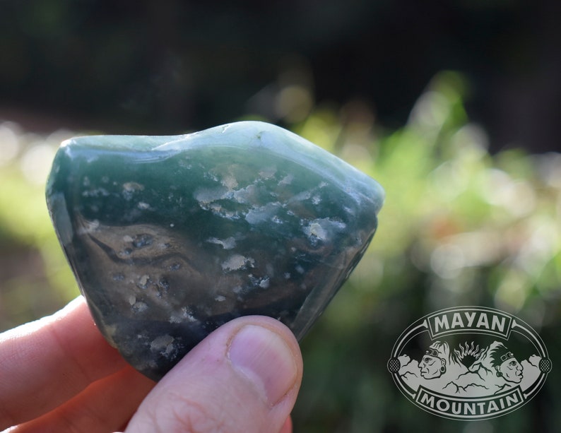 May include: A green and white agate stone with a rough, natural texture. The stone is held in a hand, and the background is blurred.