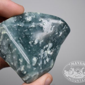 May include: A large, light blue, green, and white, irregularly shaped, polished stone with a white vein running through it.  The stone is being held in a hand.  The image has a watermark in the lower right corner that says "MAYAN MOUNTAIN".