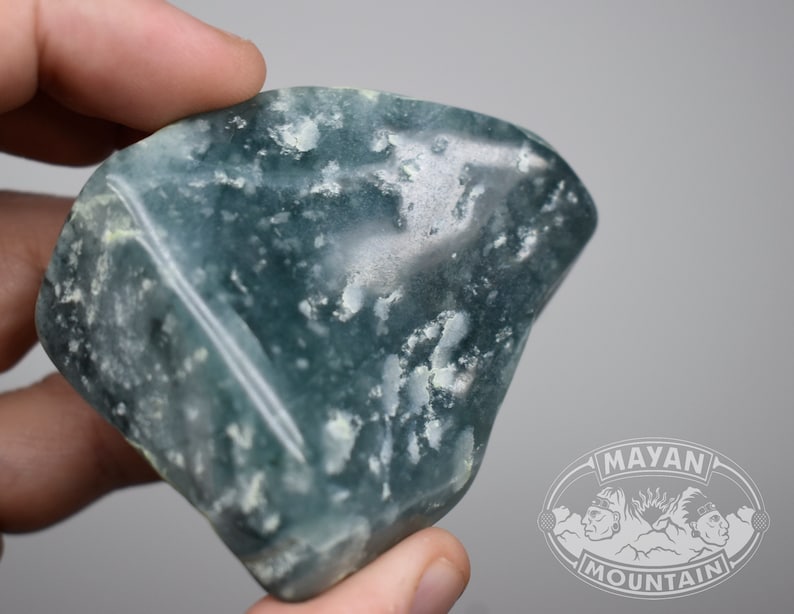 May include: A large, light blue, green, and white, irregularly shaped, polished stone with a white vein running through it.  The stone is being held in a hand.  The image has a watermark in the lower right corner that says "MAYAN MOUNTAIN".