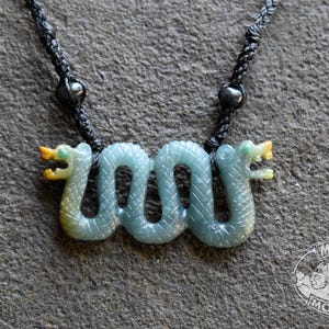May include: A pendant featuring a carved light green serpentine stone shaped like a double-headed snake with textured scales and yellow accents on the heads. The pendant is suspended from a black braided cord with two small black beads.