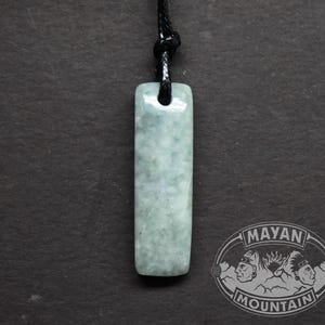 Puede incluir: Un colgante rectangular de jade verde pálido cuelga de un cordón negro. El colgante tiene una superficie lisa y pulida con sutiles variaciones de color. El cordón está anudado en la parte superior y el colgante está centrado sobre un fondo oscuro. El logotipo de Mayan Mountain está en la esquina inferior derecha.