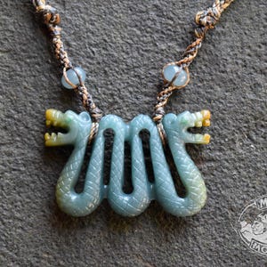 May include: A carved blue-green stone pendant featuring two serpent-like figures with yellow-tipped snouts, suspended from a braided brown cord with two small light blue beads. The logo MAYAN MOUNTAIN is visible in the bottom right corner.