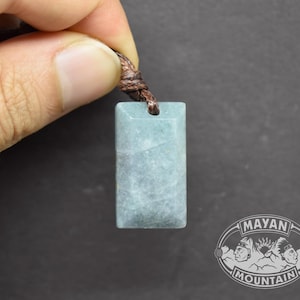Puede incluir: Un colgante rectangular azul claro con un cordón marrón. El colgante tiene un aspecto veteado y está sujeto por una mano. El logotipo de Mayan Mountain es visible en la esquina inferior derecha.