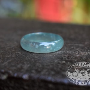 May include: A smooth, translucent light blue ring with a polished finish. The band has a rounded profile and a consistent thickness throughout. A circular logo featuring the text MAYAN MOUNTAIN and two profile faces is visible in the corner.