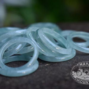 May include: A collection of smooth, pale mint-green jade rings with a polished finish. The rings are stacked together in a cluster. A logo in the bottom right corner features the text MAYAN MOUNTAIN with two profile faces and a sunburst design.